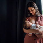 Mother breastfeeding baby boy while standing by window at home File name: