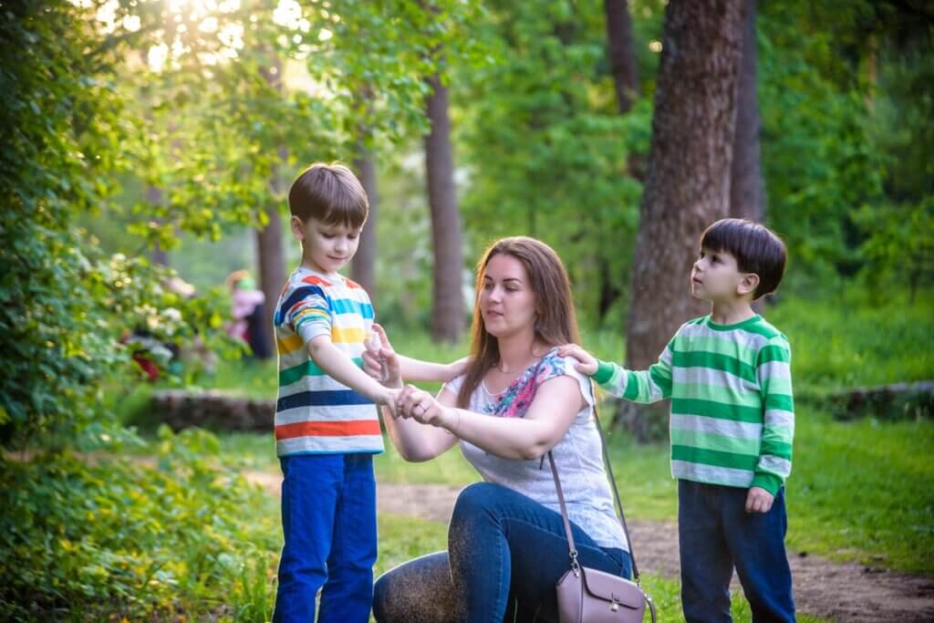 Young woman mother applying insect repellent to her two son before forest hike beautiful summer day or evening. Protecting children from biting insects at summer. Active leisure with kids.