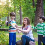 Young woman mother applying insect repellent to her two son before forest hike beautiful summer day or evening. Protecting children from biting insects at summer. Active leisure with kids.