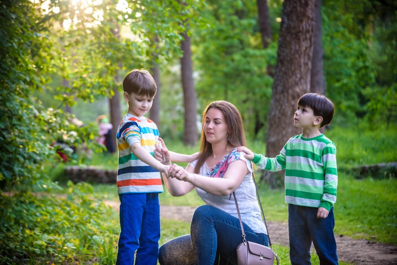 Young woman mother applying insect repellent to her two son before forest hike beautiful summer day or evening. Protecting children from biting insects at summer. Active leisure with kids.