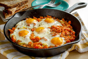 Fried eggs and sweet potato hash in cast iron skillet sitting on yellow striped kitchen towel with whole grain toast and stacked plates - healthy breakfast ideas for fall