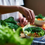 Female hand pours green onions in a bowl with green peas, cucumbers, carrots, lettuce and dill standing on a table as an example of how a dietitian can help you meet your health goals
