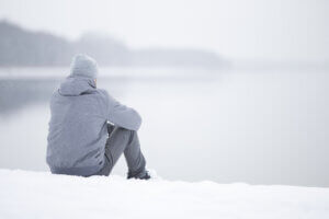 Young adult man sitting alone on snow at lake shore and looking far away. White cold snowy winter day. Thinking about life. 