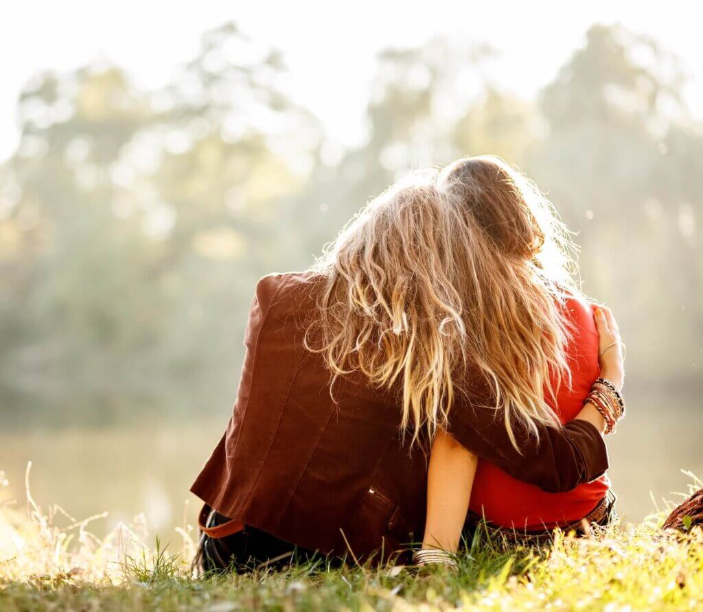 two young women sitting on grass hugging rear view