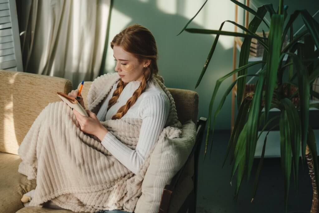 Ginger woman writing ideas in notebook, doing beauty face mask for skincare sitting in living room at home. Creative female looking thoughtful while planning routine in notebook self care daily routine