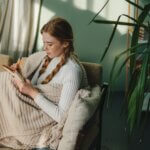 Ginger woman writing ideas in notebook, doing beauty face mask for skincare sitting in living room at home. Creative female looking thoughtful while planning routine in notebook self care daily routine