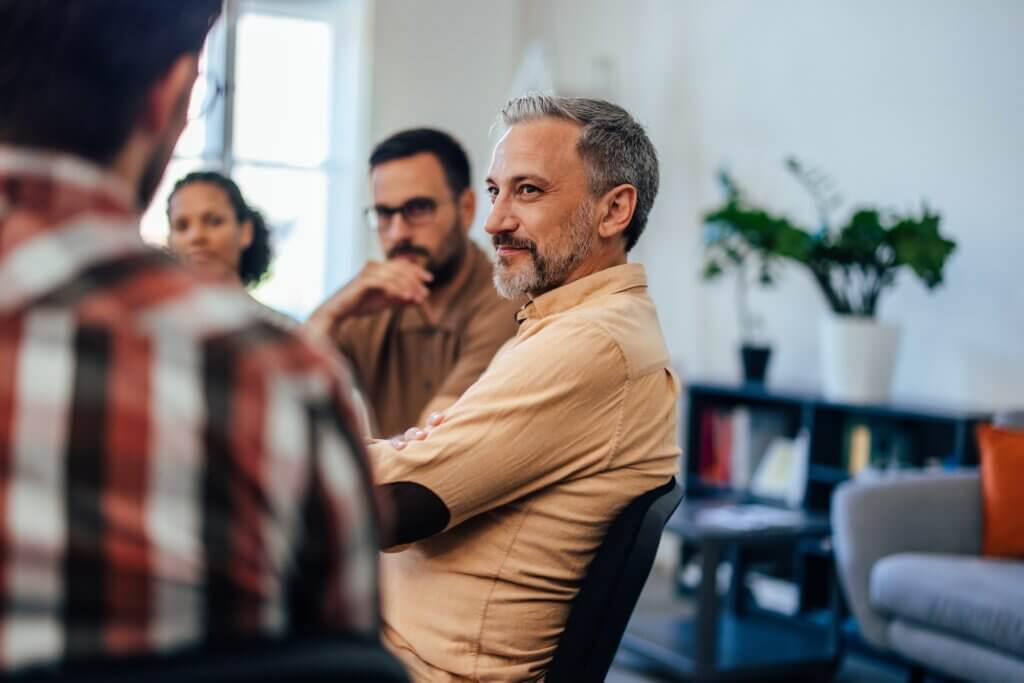 People sit in a circle, attend group therapy, and focus on the mature man.
