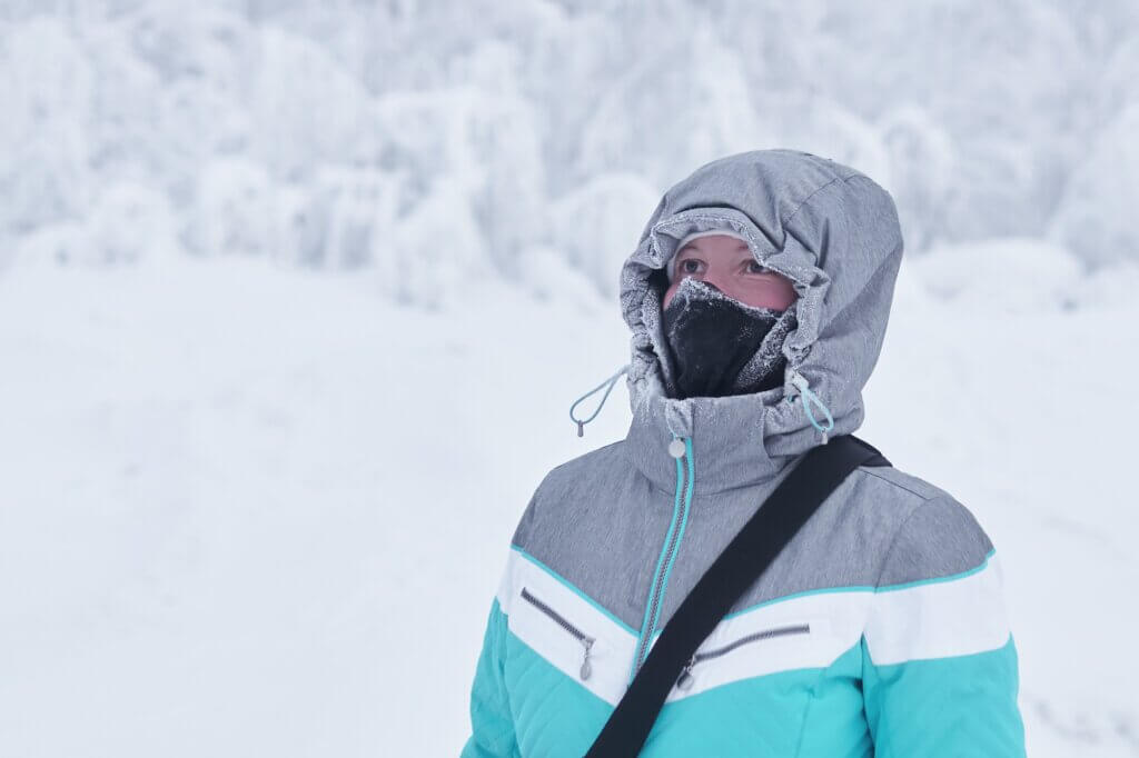 young woman in a warm jacket with a hood and a buff neck warmer on her face against the background of blurred winter landscape