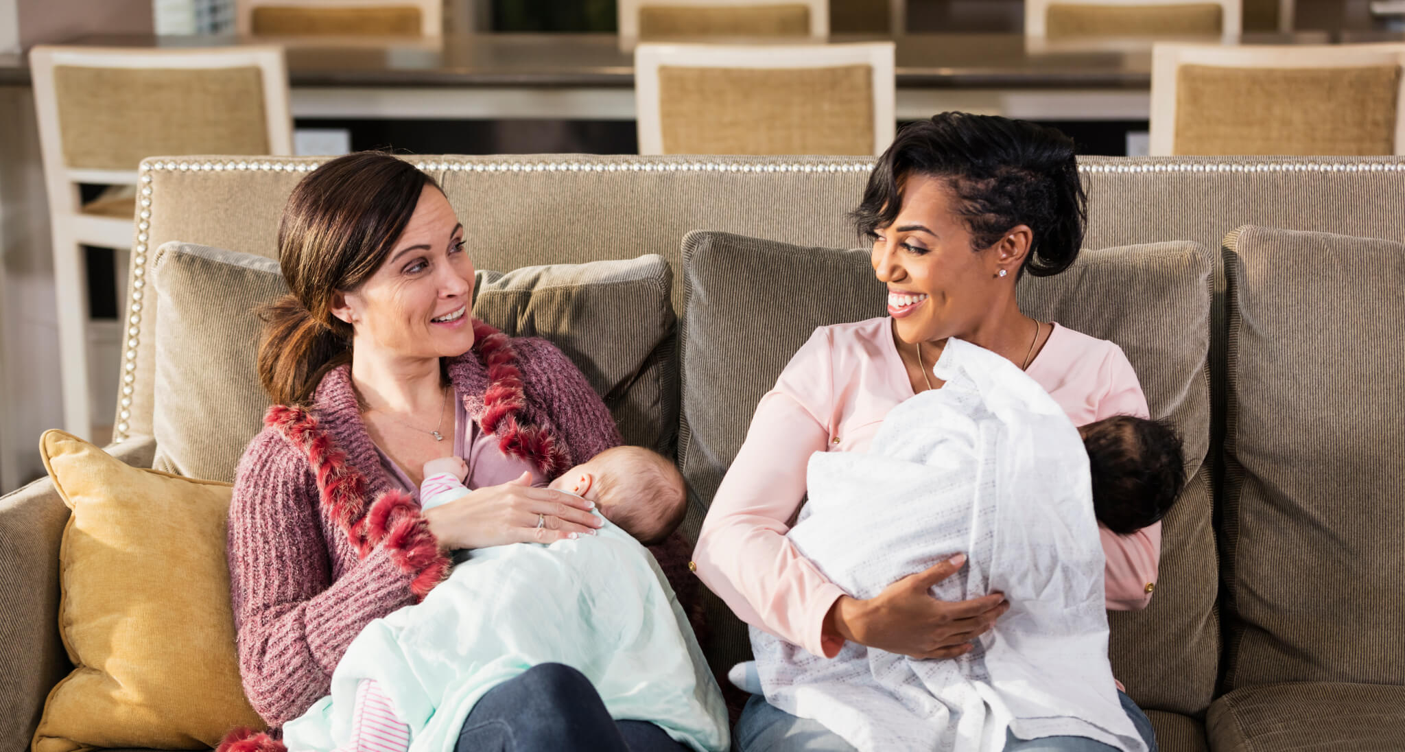 Two mothers offer each other support while breastfeeding their newborns on a couch.