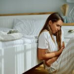 Young woman with stomach pain sitting on the floor in bedroom. Early Signs of PCOS