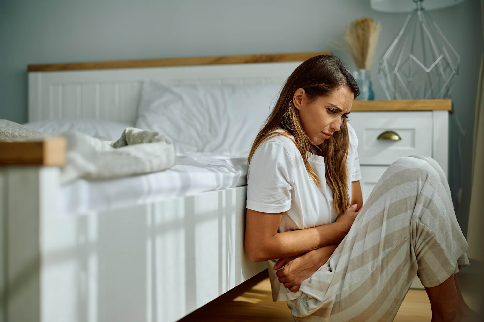 Young woman with stomach pain sitting on the floor in bedroom. Early Signs of PCOS