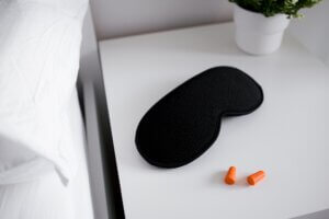Black sleep mask and orange earplugs on white nightstand with green plant. sleep Hygiene Tips