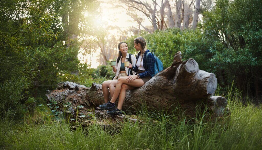 Relax, coffee and hiking with friends in nature for lunch break, trekking adventure and park conservation. Hungry, conversation and environment with women on tree in forest for snack and peace. healthy hiking snacks