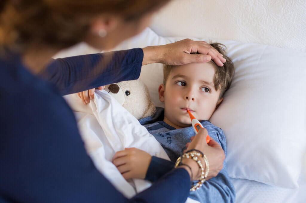 Sick boy with thermometer laying in bed and mother hand taking temperature. Mother checking temperature of her sick son who has thermometer in his mouth. Sick child with fever and illness while resting in bed. what is measles featured image