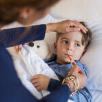 Sick boy with thermometer laying in bed and mother hand taking temperature. Mother checking temperature of her sick son who has thermometer in his mouth. Sick child with fever and illness while resting in bed. what is measles featured image