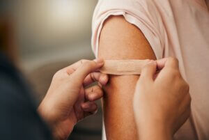 Covid, vaccine and plaster with a doctor and patient consulting in an appointment or checkup at home. Nurse, healthcare and trust with a health professional using a bandaid on an arm after medication.