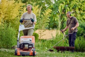Male gardener trimming the lawn with a grass trimmer while his colleague uses a lawnmower