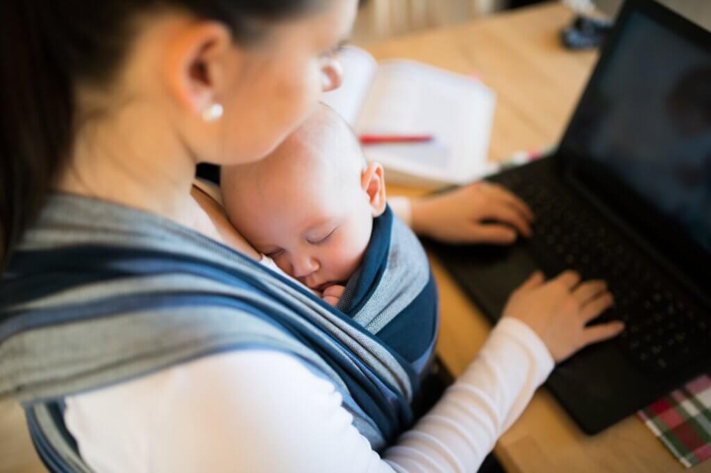 Working mother with her newborn baby son sleeping in sling at home, sitting at the table, writing on notebook keyboard breastfeeding at work laws featured image