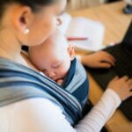 Working mother with her newborn baby son sleeping in sling at home, sitting at the table, writing on notebook keyboard breastfeeding at work laws featured image