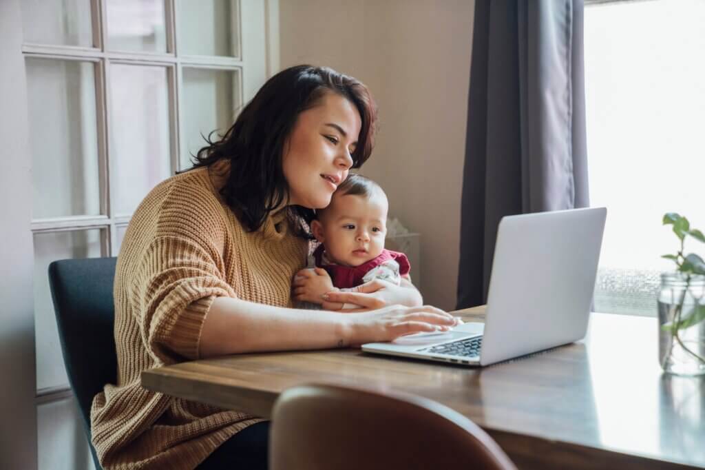 Mom holding baby and working from home on laptop at dining room table