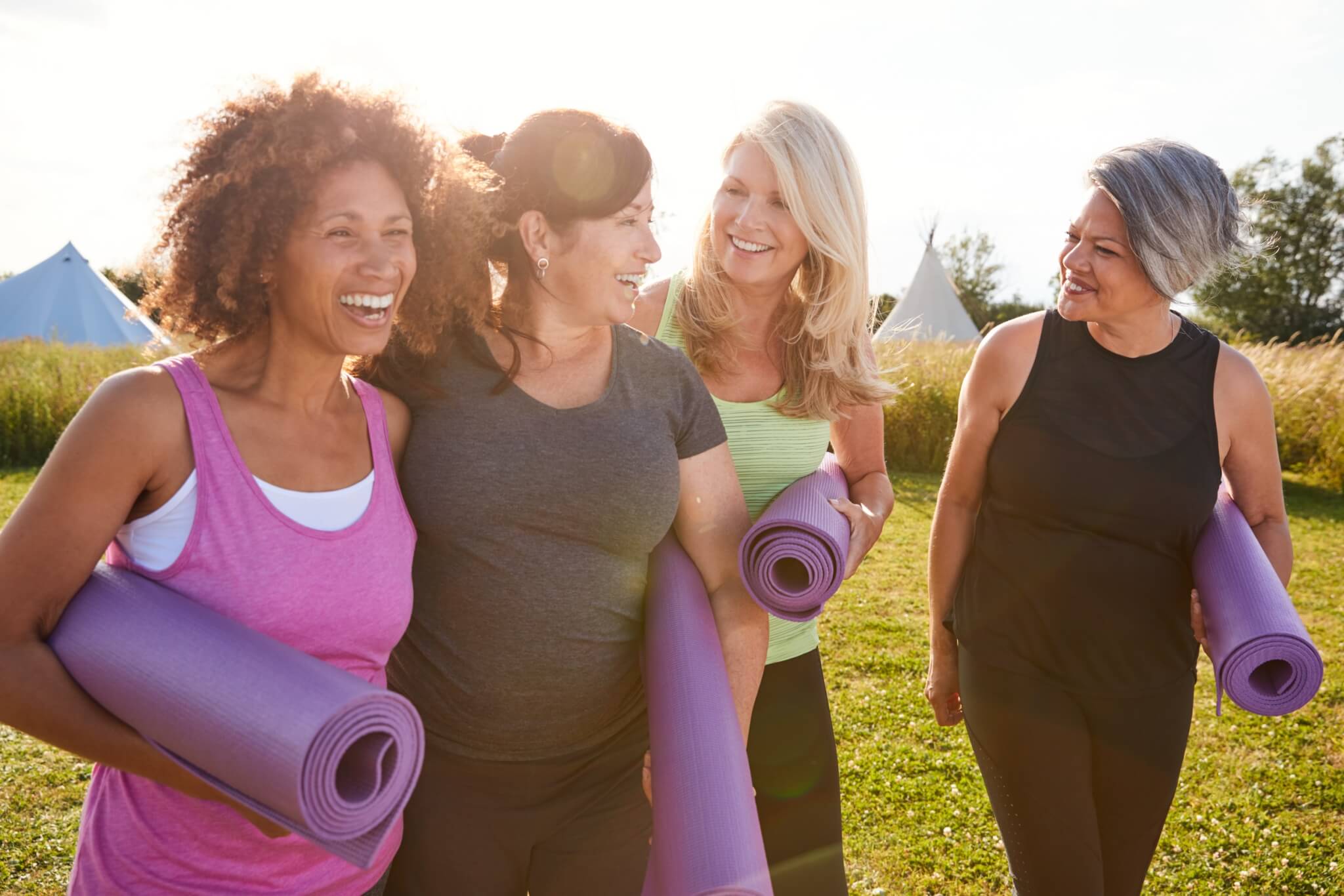 four middle-aged women walking together while holding yoga mats on a warm sunny day