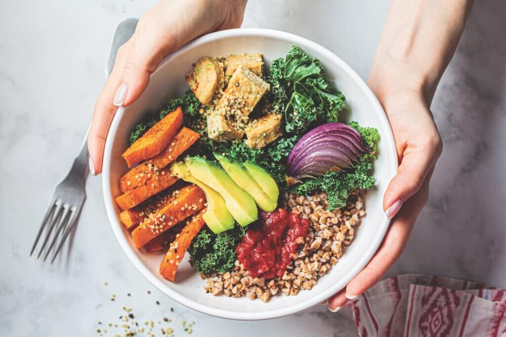 hands holding a bowl of high fiber gut microbiome friendly foods in a bowl, including tofu, kale, faro, red onion, avocado, and sweet potato