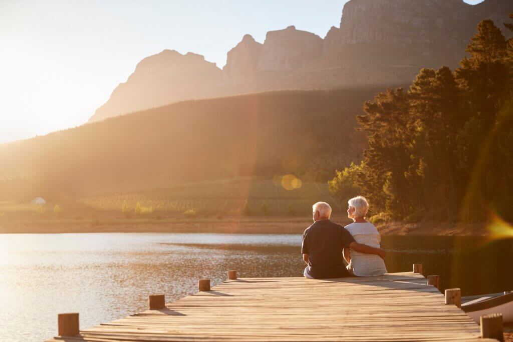 a senior couple sitting on the edge of a wooden dock on a lake surrounded by mountains while warm golden light pours across the scene