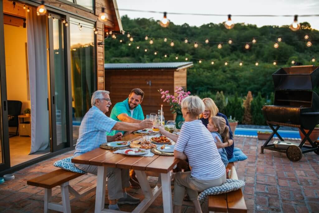 three generations of people sitting at an al fresco dining table with bistro lights strung above them as they enjoy their dinner