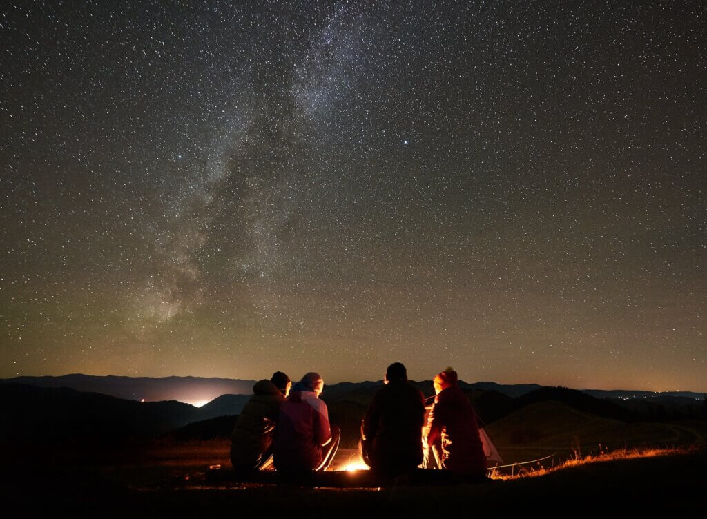 group of people sitting around a campfire with the milky way and sky full of stars above them