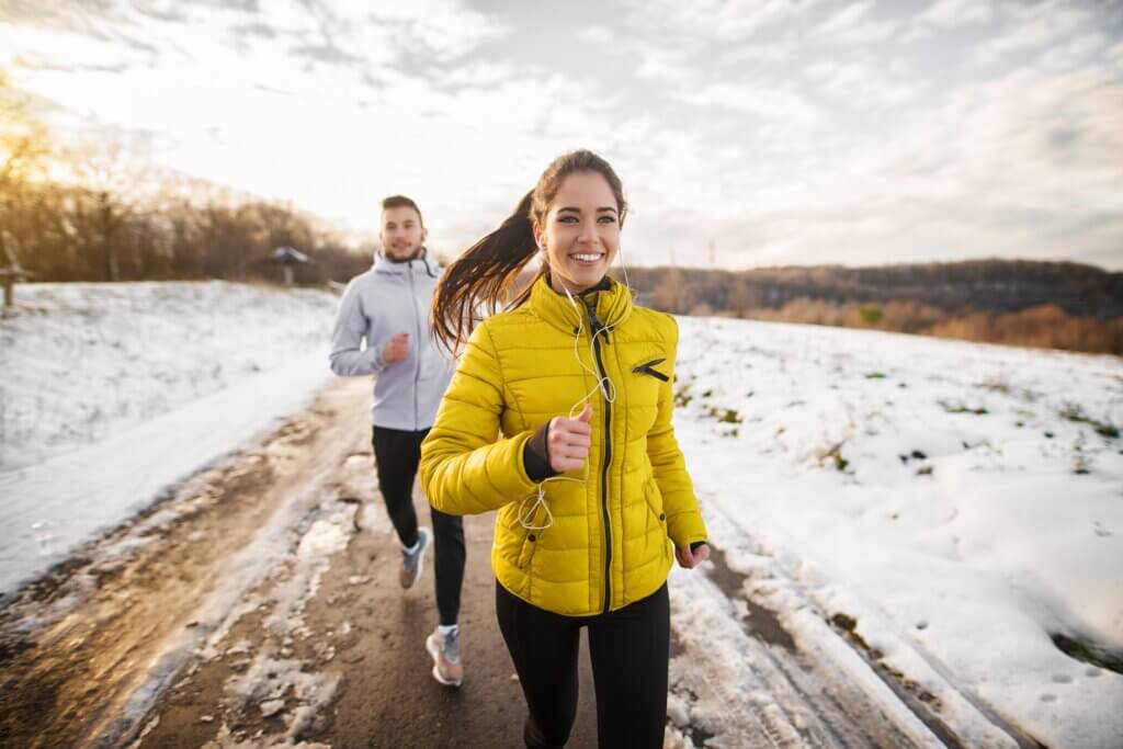 woman in a yellow puffer jacket running down a dirt road on a snowy day while a man jogs behind her