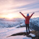 woman who does light therapy in the winter to deal with her anxiety and depression standing at the top of a snowy mountain peak looking out at the sunrise